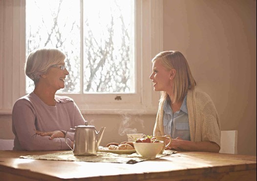 Two women sit at a table with a teapot, pastries, and a bowl of fruit, conversing near a window with bare trees visible outside.