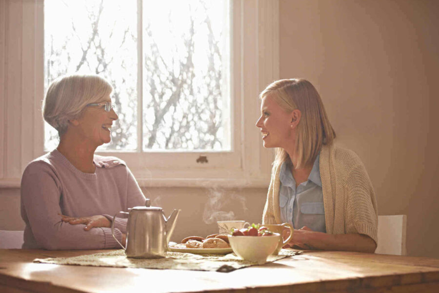 Two women sit at a table having a conversation, with a teapot, plate of pastries, and bowl of fruit in front of them. Sunlight streams in through a window behind them.