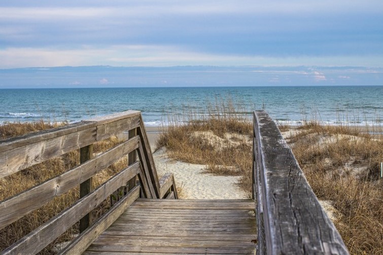 A wooden boardwalk leads over sand dunes to a beach with sea oats and gentle ocean waves under a partly cloudy sky.