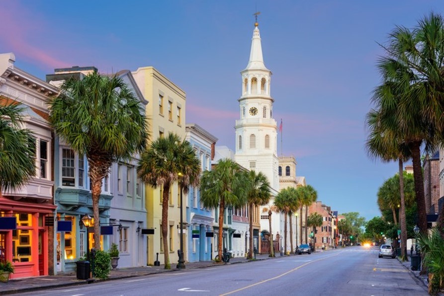 A quiet street lined with colorful buildings and palm trees, featuring a white church with a tall steeple under a pastel evening sky.