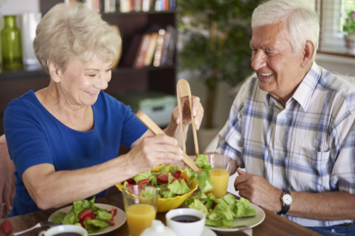 Two older adults are sitting at a dining table, smiling, and serving salad. There are glasses of orange juice and a bowl of salad on the table in front of them.