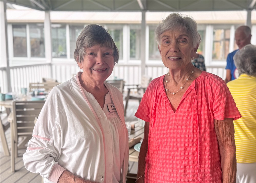 Two older women stand side by side indoors, one wearing a white shirt and name tag, the other in a pink top. Tables and chairs are visible in the background.