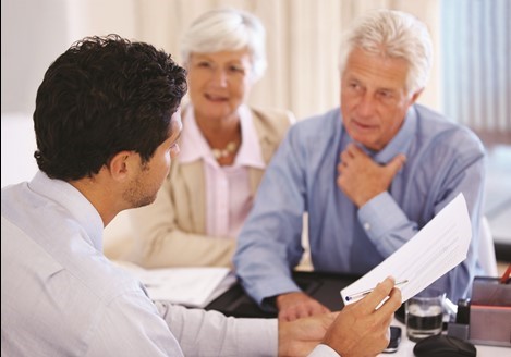A man in a suit shows documents to an older couple seated at a desk, engaged in a conversation.