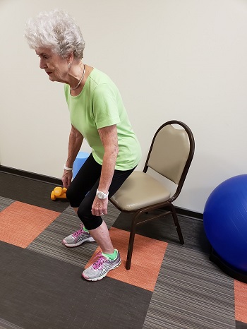 Older woman in athletic wear performs a sit-to-stand exercise from a chair in a fitness room with an exercise ball and dumbbell nearby.