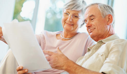Two older adults sit together on a couch, looking at a document and discussing its contents in a well-lit room.