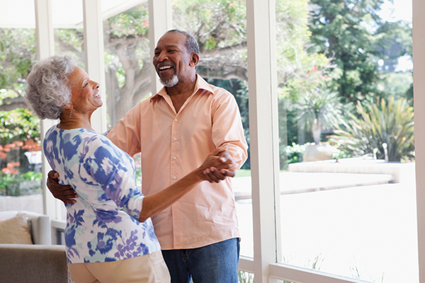 Two older adults smile and dance together in a bright room with large windows, with trees and greenery visible outside.