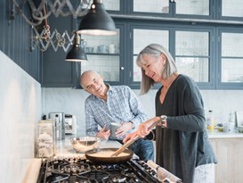 An older woman cooks at a stove while an older man smiles and watches from the kitchen counter in a modern home kitchen.