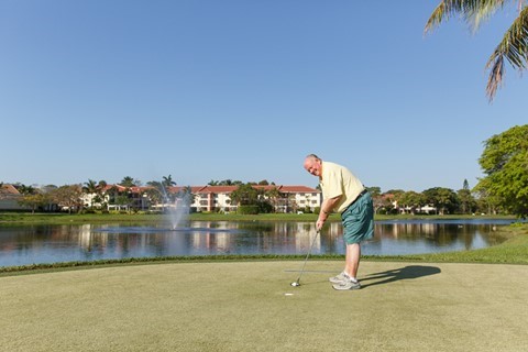 A man stands on a golf green, putting a ball toward the hole, with a pond, fountain, palm trees, and residential buildings in the background under a clear blue sky.