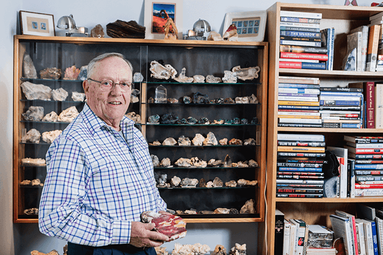 An older man stands smiling in front of a display case filled with rocks and minerals, holding a specimen, with shelves of books and more rocks in the background.
