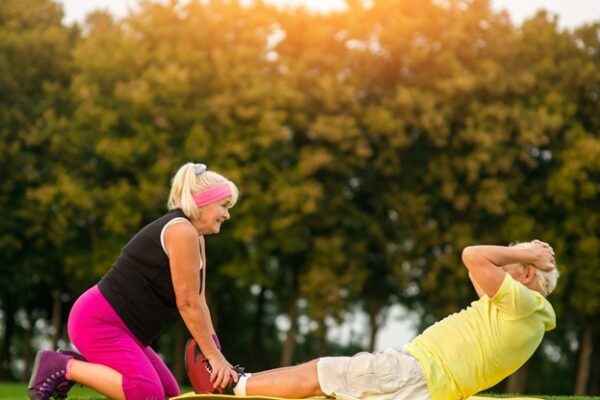 An older woman holds an older man’s feet as he does sit-ups on a mat outside in a grassy area with trees in the background.