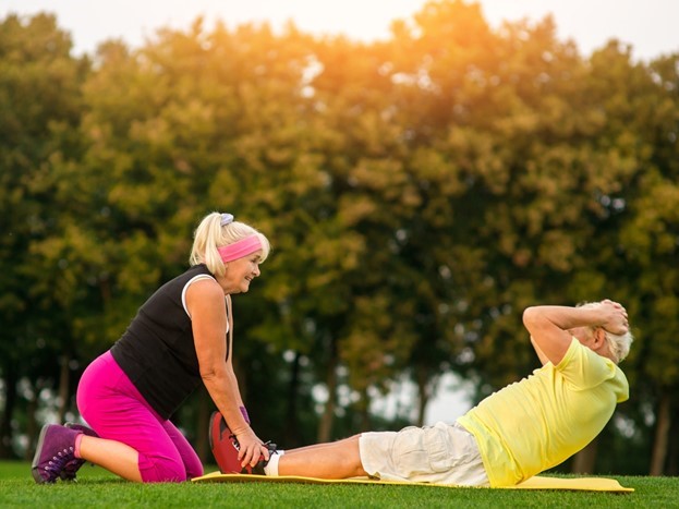 An older woman holds an older man’s feet as he does sit-ups on a mat outside in a grassy area with trees in the background.