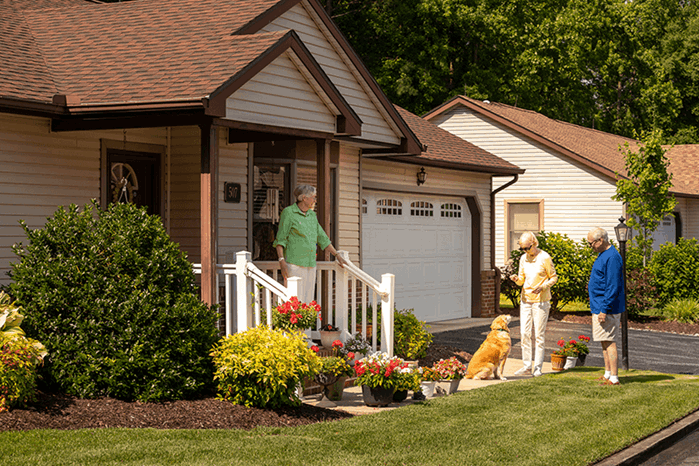 Three people and a dog converse outside near the porch of a house surrounded by plants and flowers on a sunny day.