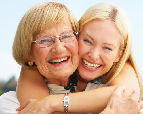 An older woman and a younger woman smile and embrace outdoors, both looking at the camera.