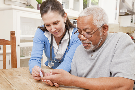 A healthcare professional helps an older man check his blood sugar at a table in a kitchen setting.