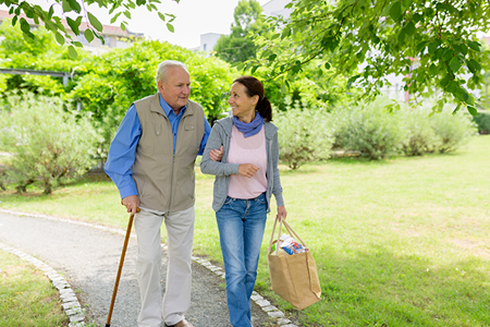 An older man with a cane walks arm-in-arm with a woman carrying a grocery bag along a park path, surrounded by greenery.