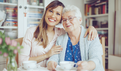 Young woman and elderly woman sit together at a table, smiling at the camera, with cups and bookshelves in the background.
