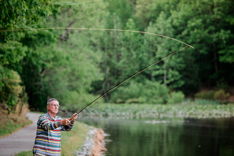 An older man in glasses stands near a pond, casting a fishing rod. Green trees and lily pads surround the calm water.