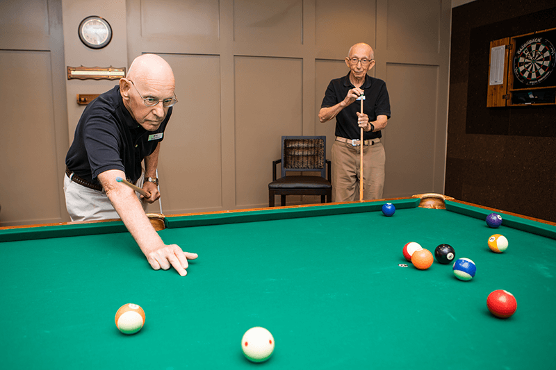 Two elderly men play pool in a game room; one lines up a shot while the other stands nearby holding a cue stick.