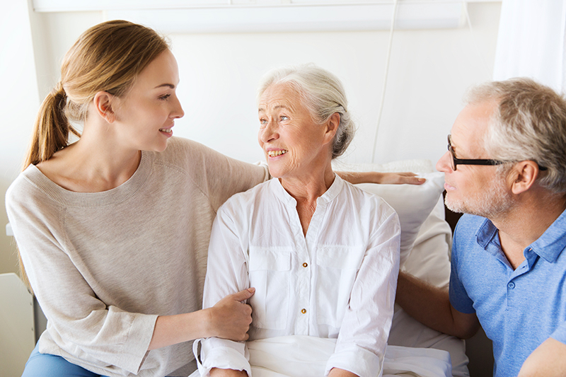 Young woman and older man sitting beside an older woman in a hospital bed, all engaging in conversation and showing support.