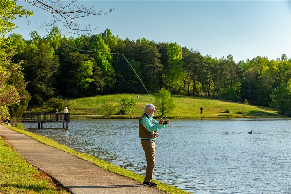 An older man stands on a lakeshore fly fishing, with a wooded area and two people on a dock in the background.