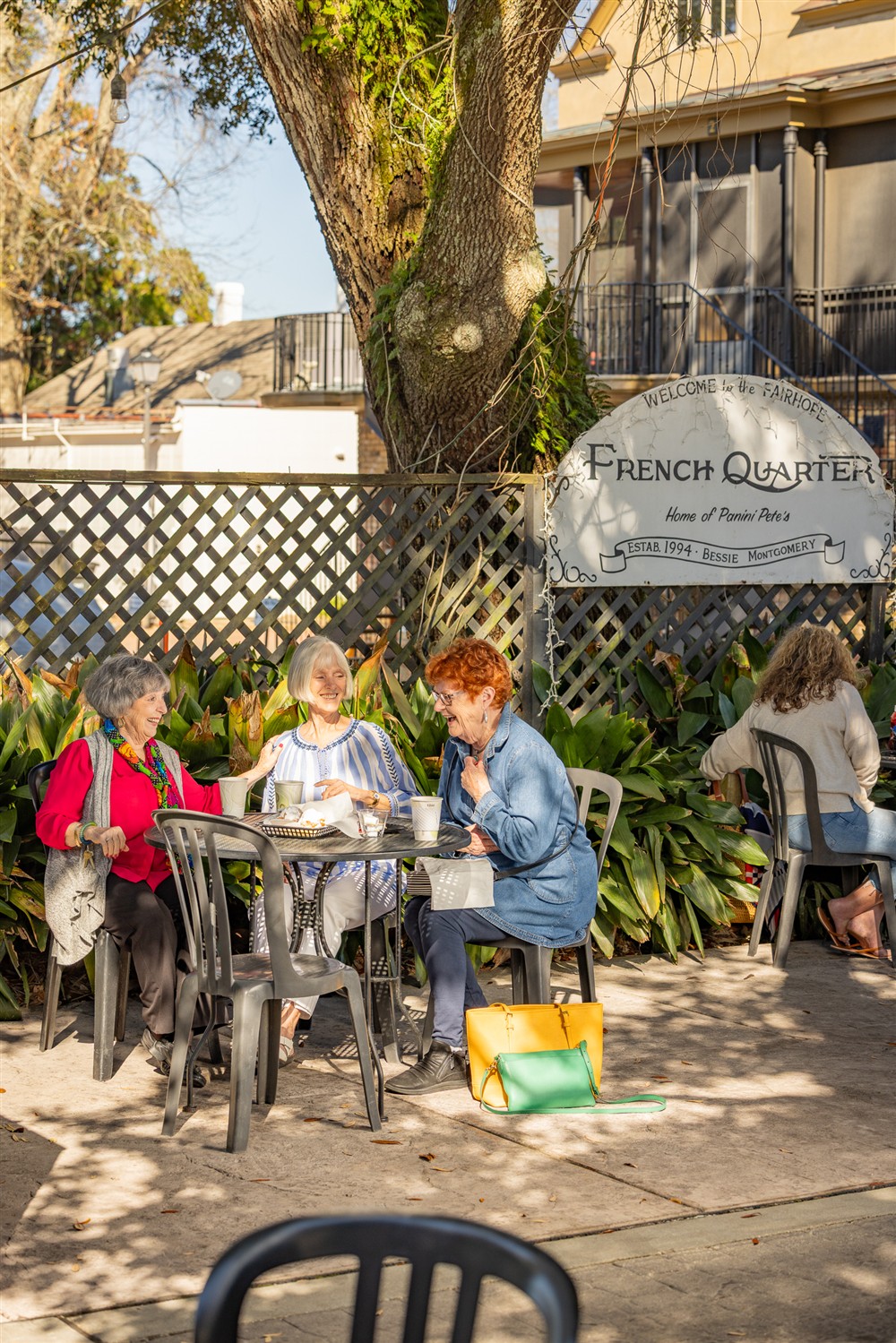 Three women sit at an outdoor café table under a tree, eating and talking. A sign behind them reads “French Quarter Home of Beignet Bites.” Another person sits alone nearby.