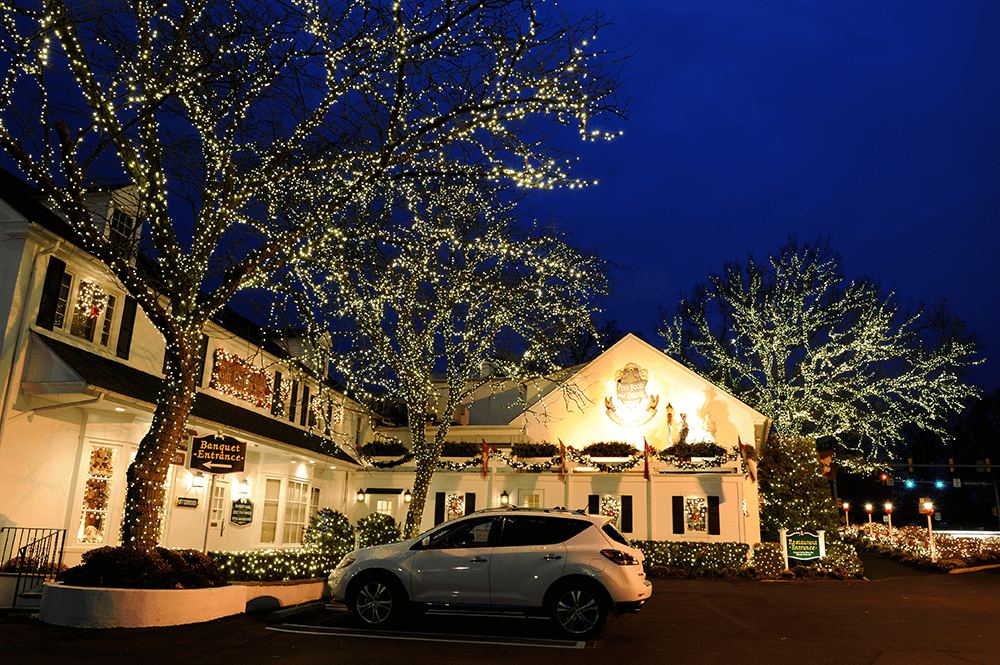A white car is parked outside a building decorated with white holiday lights on trees and the facade at night.