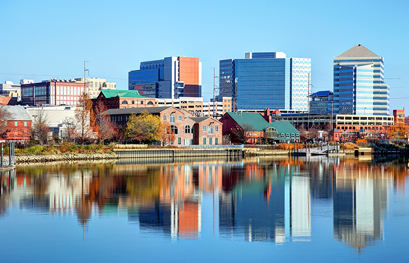 Skyline of a city with modern and historic buildings reflected in a calm river under a clear blue sky.