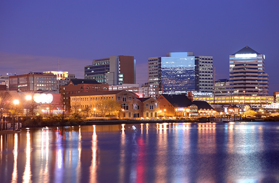 City skyline at dusk with office buildings and waterfront reflecting lights in calm water.