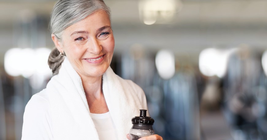 Older woman with gray hair and a white towel around her neck smiles while holding a water bottle in a brightly lit gym or fitness center.