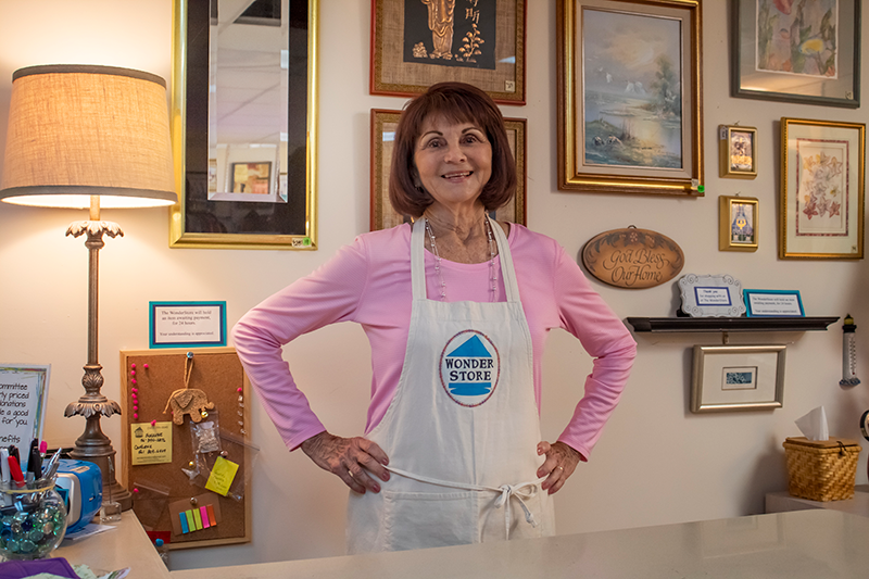 A woman in a pink shirt and apron stands behind a counter in a store, surrounded by framed artwork and various decorations on the wall.