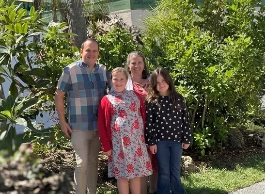 A man, a woman, and two girls posing together outdoors in front of a green hedge.