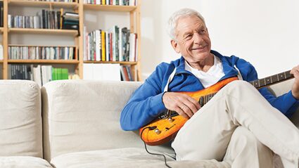 An older man in a blue hoodie sitting on a sofa and playing an electric guitar.